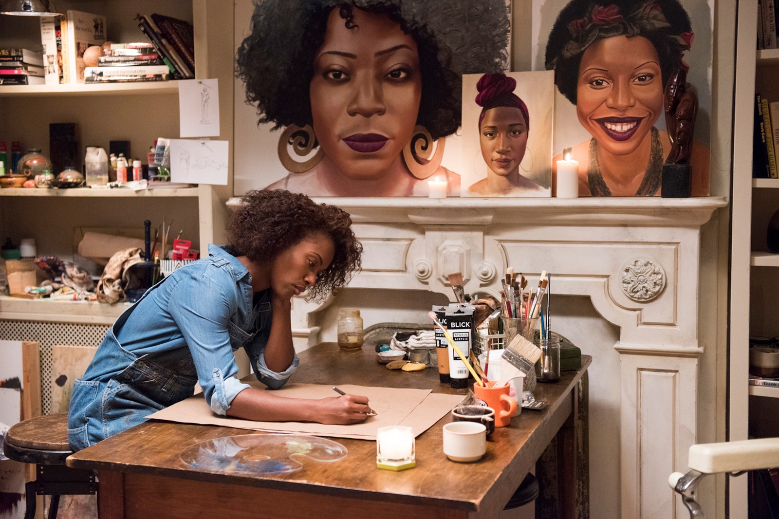 Nola desenhando numa mesa, repleta de ferramentas de pintura. Ao fundo, alguns de seus desenhos de rostos de mulheres negras