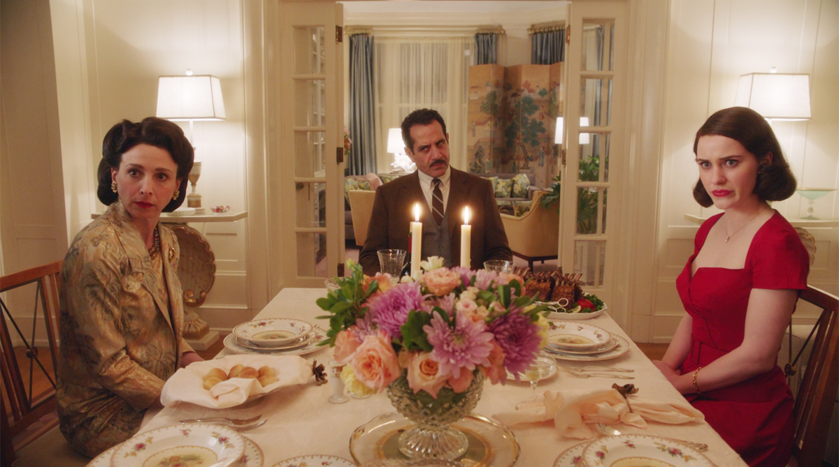 5 - Uma sala de jantar chique, com a mesa arrumada, com flores, com velas acesas e três pessoas sentadas nas cadeiras. Mãe e filha olham para algo espantadas e o homem olha zangado. To