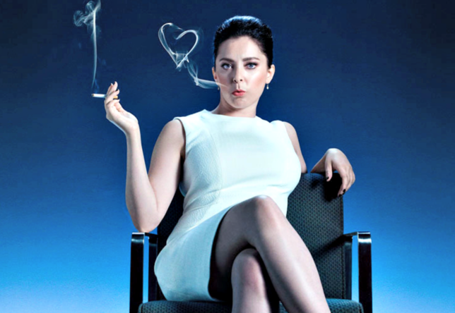 Rebecca está sentada em uma cadeira com as pernas cruzadas e usa um vestido elegante e o cabelo preso em um coque. Ela segura um cigarro e solta fumaça pela boca. A fumaça forma o formato de um coração.