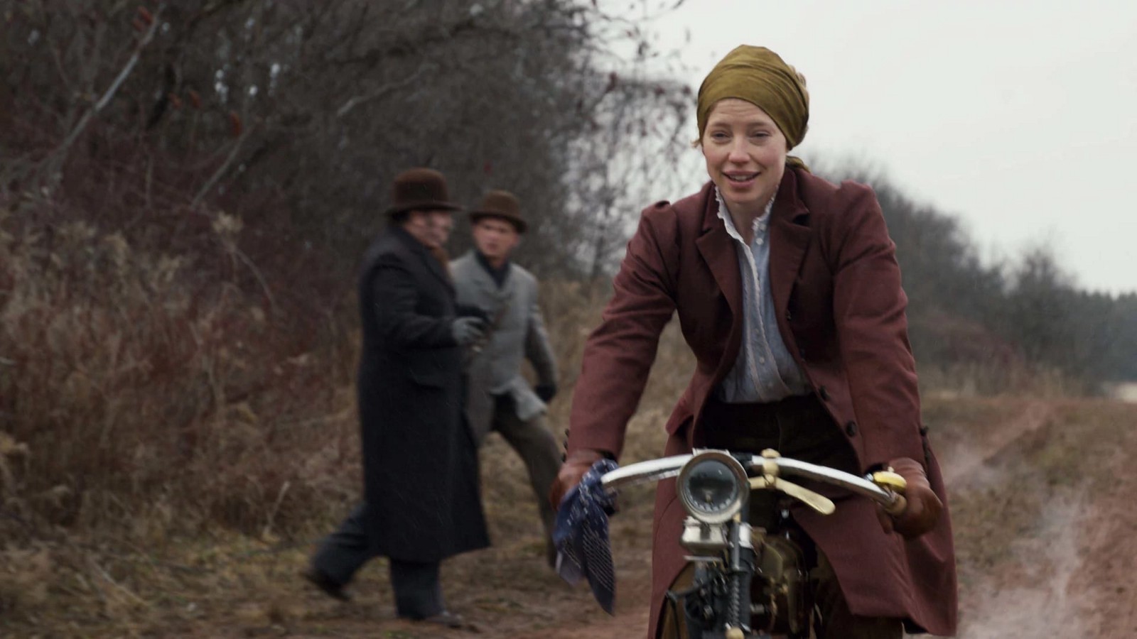 A personagem Miss Stacy, professora de Anne, pilota uma moto na estrada e dois homens olham para ela.